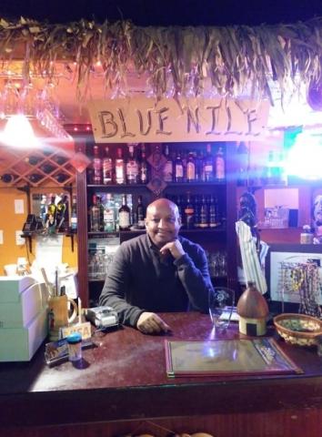 The owner of the restaurant Michael stands behind a counter decorated with a grass canopy and wooden sign that reads "Blue Nile"