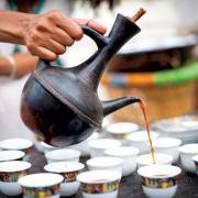 A hand pours coffee out of a tall brown ceramic pot into many small white cups